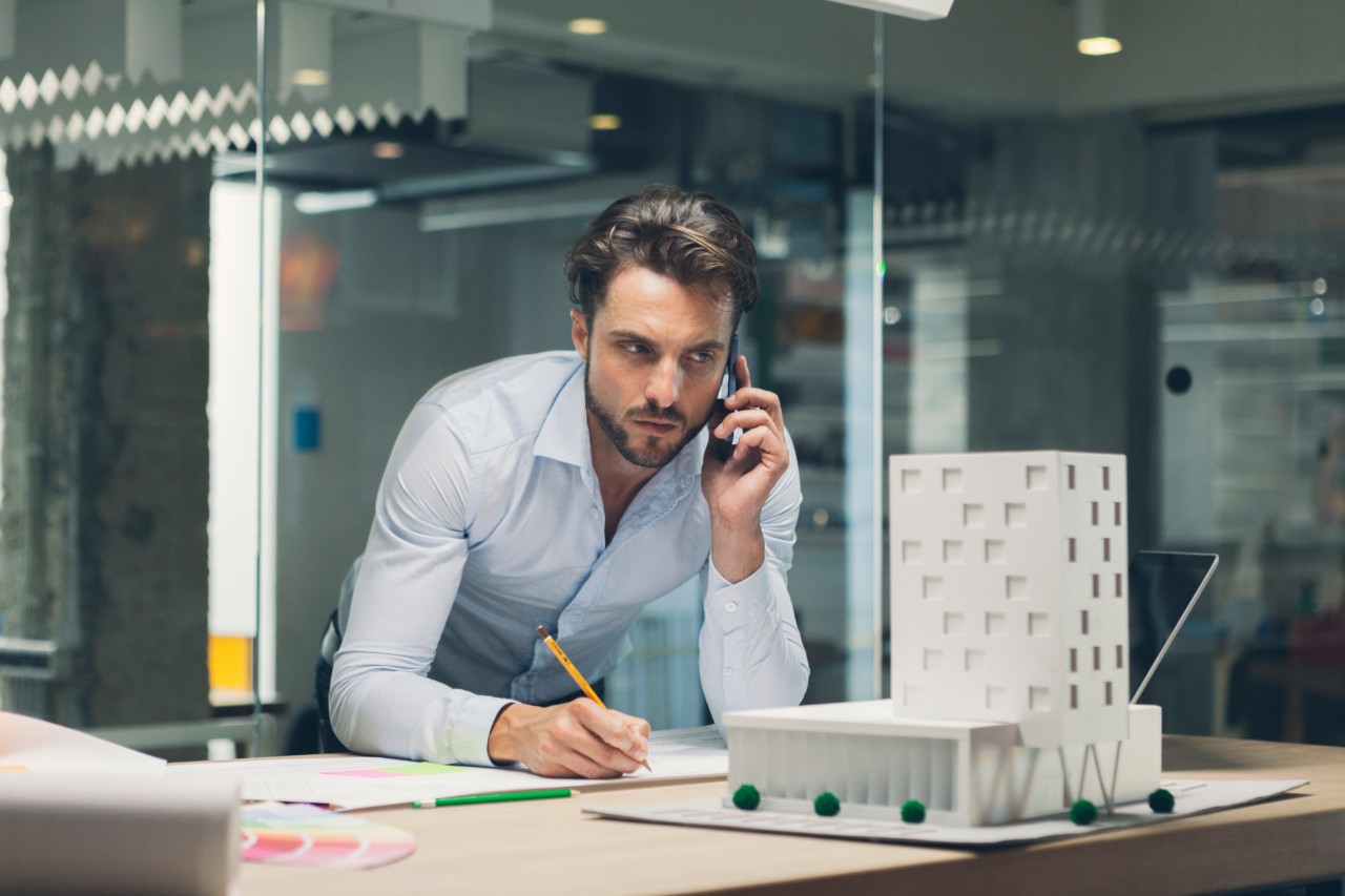 Architect working in his office. Standing and talking on smart phone and writing notes with pencil. Talking to client.